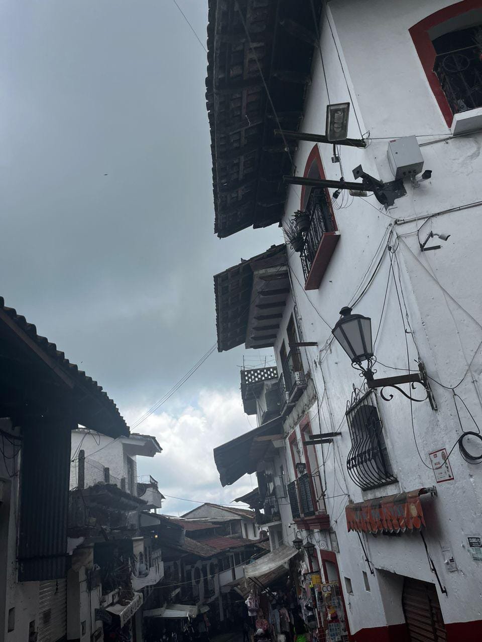 Se pueden observar construcciones antiguas altas de color blanco y muchas ventanas con tejados cafés, en ambas partes de la calle. Abajo en la imagen se aprecian algunas lonas que conforman los diferentes puestos del mercado callejero de Cuetzalan.