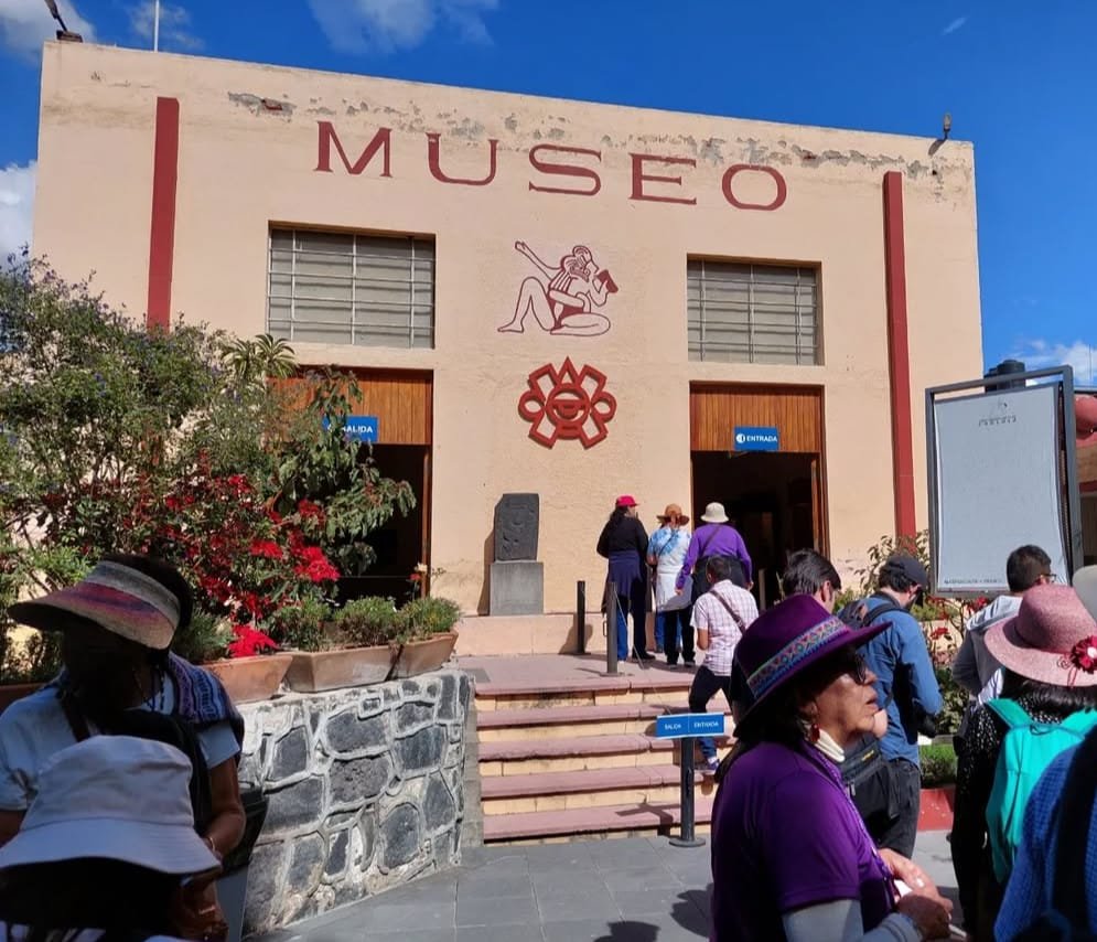 Personas aparentemente turistas, ya que la mayoría traen sombreros y mochilas, se aprecian unas flores rojas, escaleras y la entrada del museo de Sitio de Cholula, cuyo color de estructura es color naranja muy claro con toques rojos.
