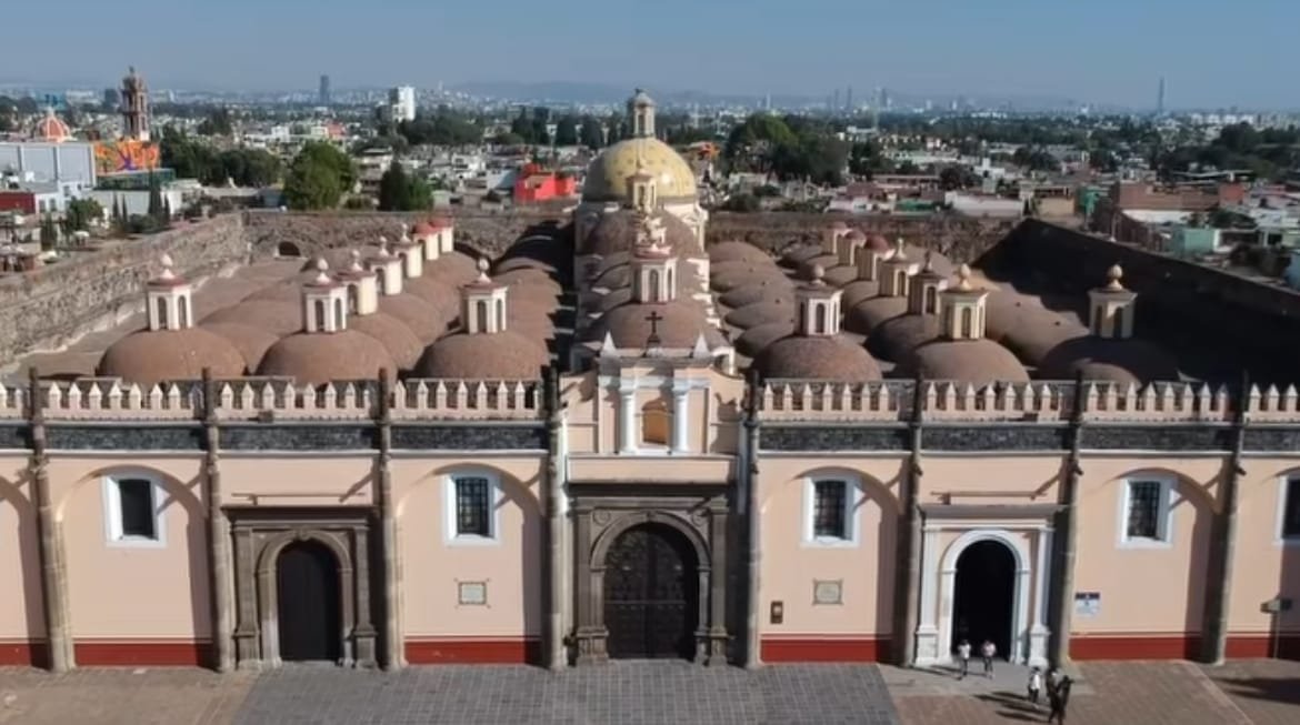 Se observa un espacio amplio con construcciones que conforman el Conjunto Convencional de San Gabriel, en donde se aprecia un patrón de techos, 3 puertas y 5 ventanas, el color de la estructura es un tono anaranjado sumamente claro, con franjas rojas en la parte inferior, al fondo se observa una parte de la ciudad con edificios, iglesias y un poco de árboles verdes.