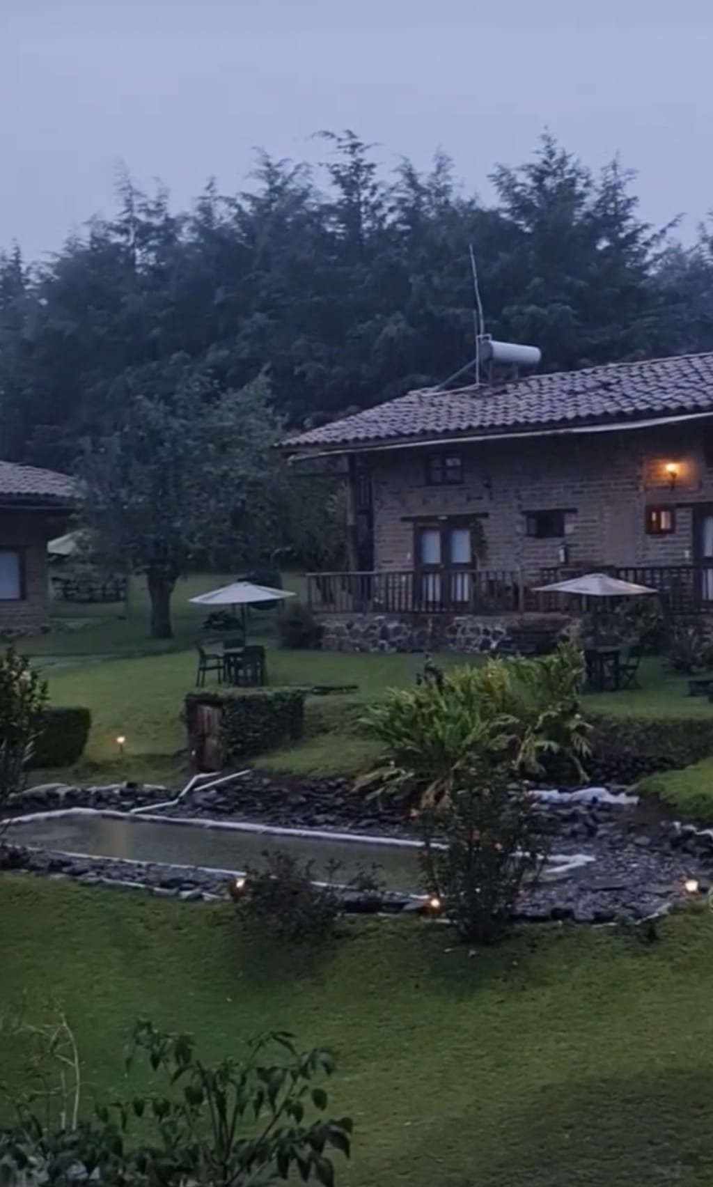 Pasto y plantas verdes por todo el lugar, una tipo de piscina o fuente decorativa, sillas y mesas con sus respectivas sombrillas y una cabaña de fondo con una estructura de piedra color café con un techo que supone ser de teja.