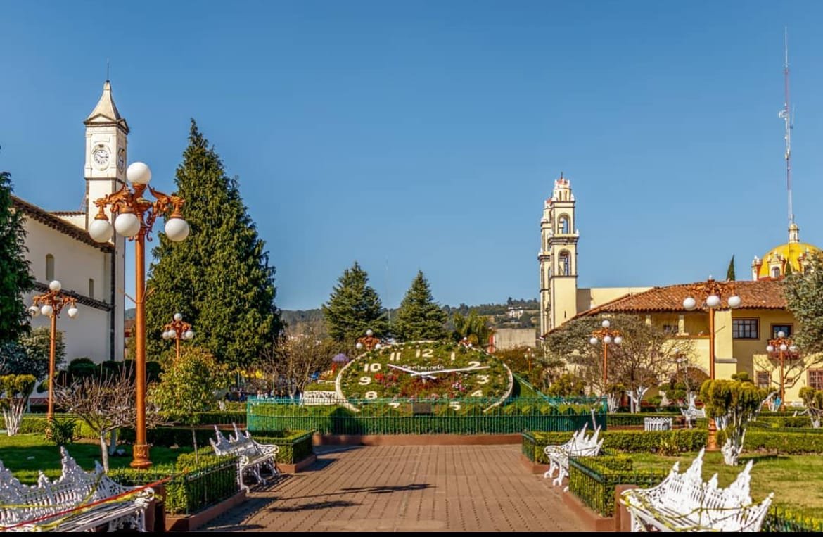 Bancas blancas con jardineras verdes a su alrededor, lámparas naranjas con focos circulares blancos, en medio se encuentra el famoso reloj constituido de plantas verdes y otros colores como rojo y blanco para los números y las manecillas. Al fondo se observan edificios blancos y amarillos con teja naranja y columnas pertenecientes a una iglesia de color blanco con detalles rojos.