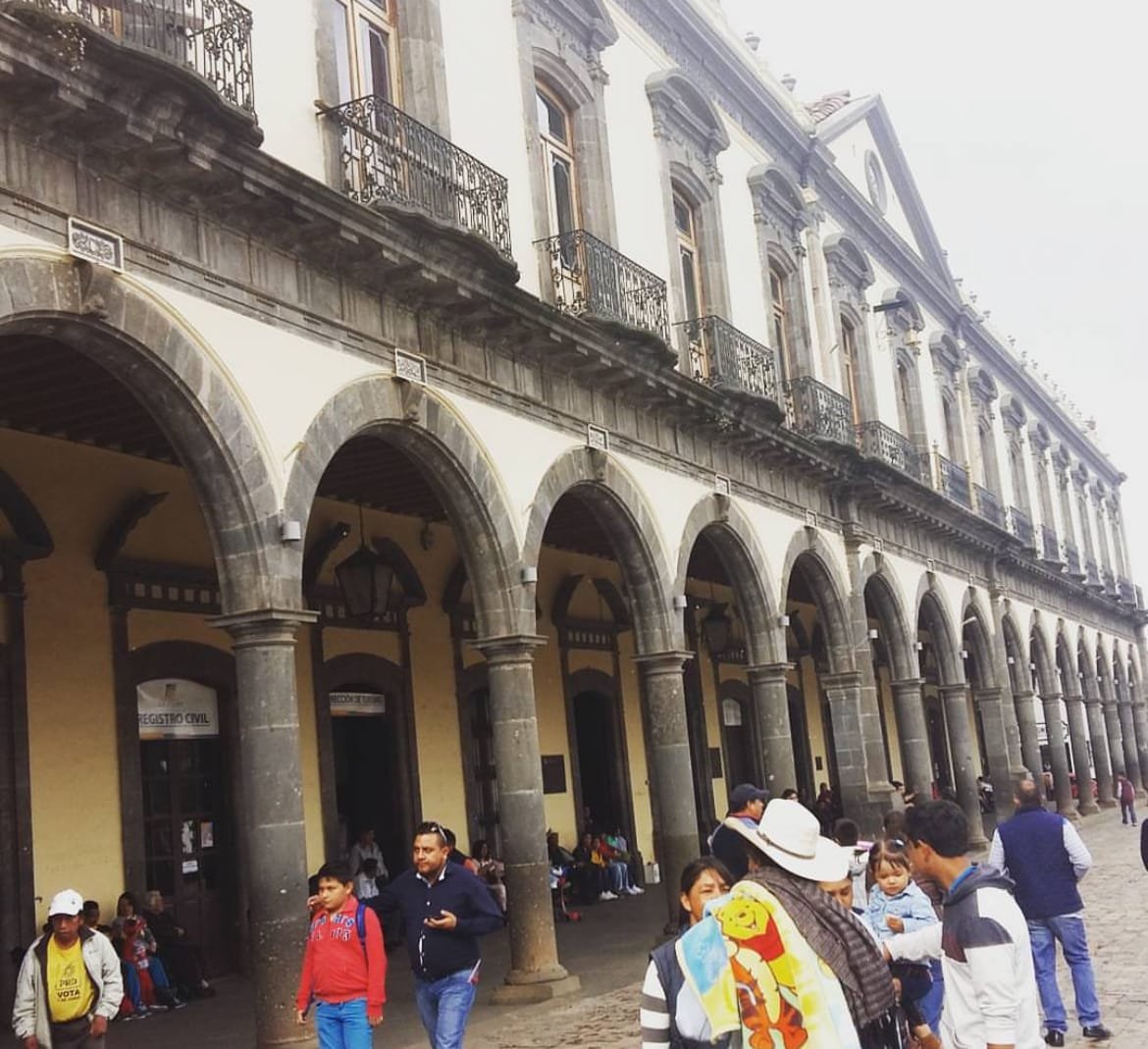 Palacio de color beige con tonos cafés con entradas grandes. A su alrededor hay varias personas caminando o conversando.
