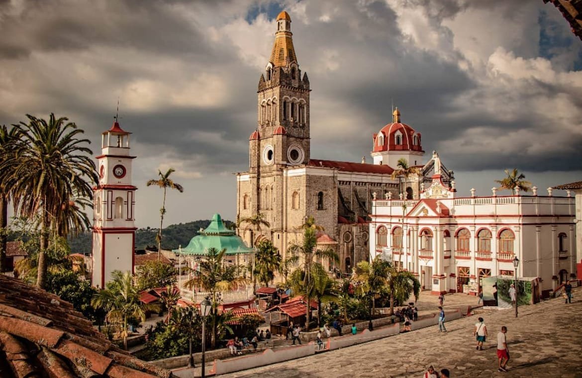 Se nota una calle de piedra, palmeras verdes, un kiosco con techo verde; cerca de este hay una torre de reloj de color blanco y rojo. Hay varias personas caminando. Al fondo se aprecia un edifico blanco y al lado la iglesia de San Francisco de Asís que tiene una torre alta.