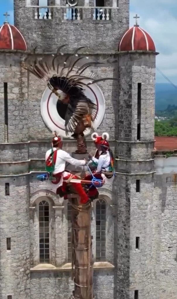 Tres hombres; uno portando un traje que hace alusión a una águila y dos abajo de él con trajes típicos de Cuetzalan. Los tres hombres se encuentran en las alturas de un tipo poste de gran altura frente a la Iglesia de San Francisco de Asís.