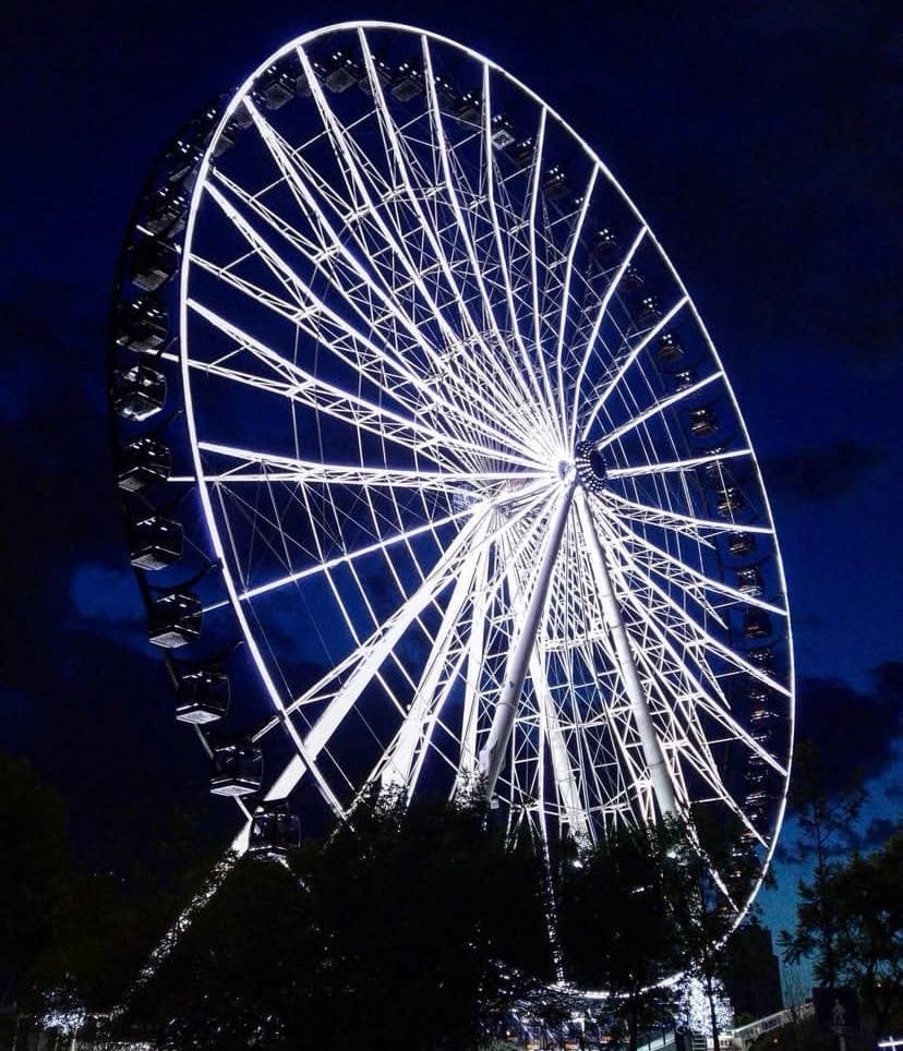 Foto tomada desde abajo de la rueda de la fortuna de Puebla, mejor conocida como la Estrella de Puebla, iluminada por luces blancas y tomada en la noche, resaltando su estructura.