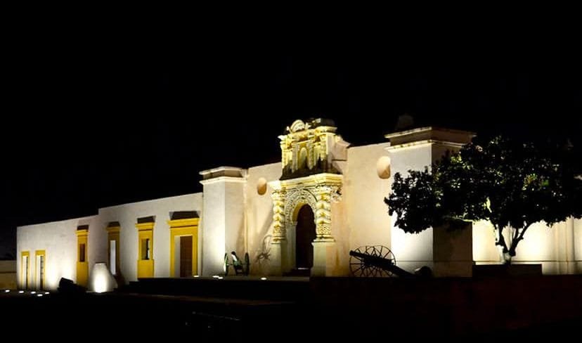 Foto tomada en la noche de la fachada de la entrada de los Fuertes de Loreto y Guadalupe, iluminado por lámparas en el piso. Se observan objetos antiguos y un árbol en el exterior.