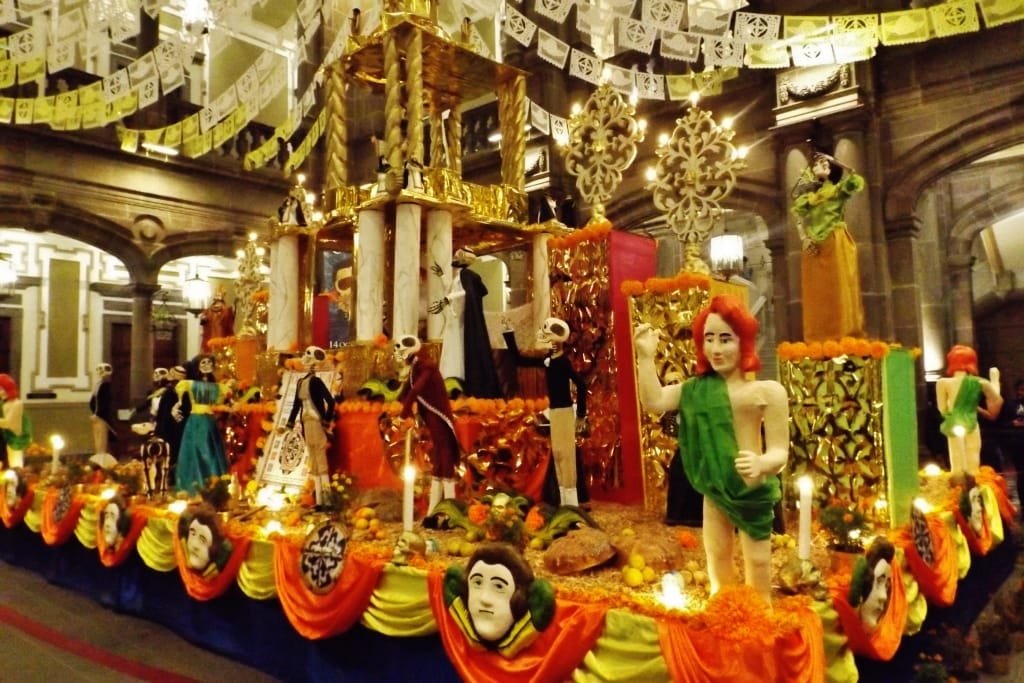 Se observa una ofrenda tradicional del Día de Muertos en la Casa de la Cultura de Puebla, decorado con papel picado, velas y figuras coloridas, incluyendo calaveras, esqueletos vestidos y esculturas humanas. El altar está adornado con flores de cempasúchil, frutas y elementos simbólicos de la cultura mexicana.